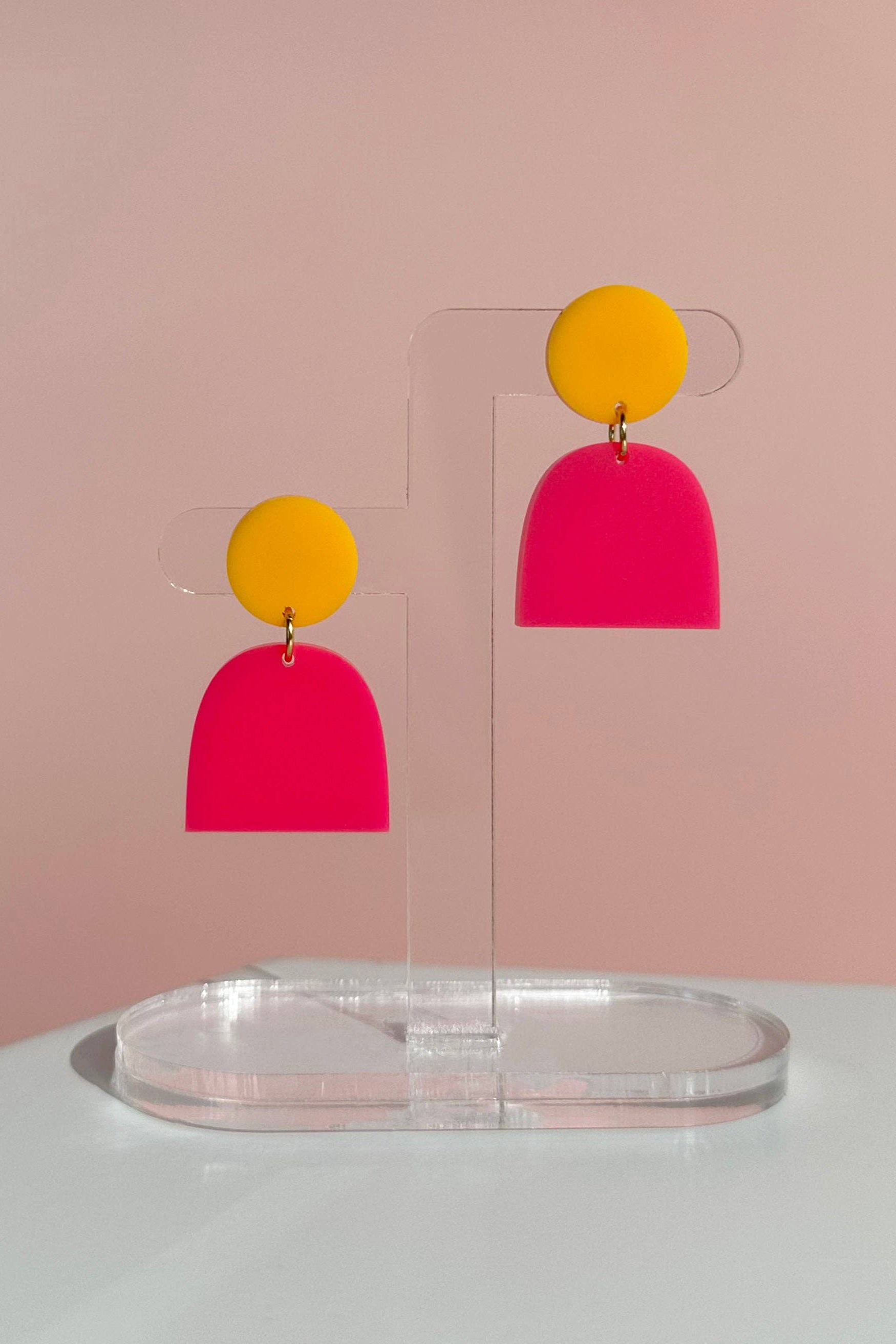 Pair of colorful earrings on a clear stand against a pink background