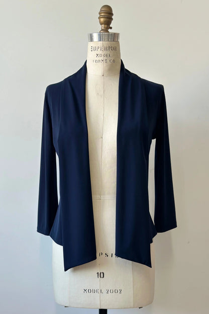 The Lioren Cardigan by Kollontai in Navy, a stretchy cropped jacket with longer pointed panels in the front that can be worn loose or knotted, is shown on a mannequin in front of a white background. 
