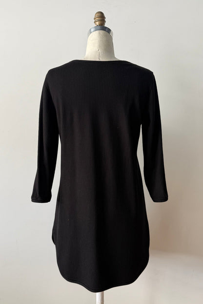 A back view of the Clara Tunic by Kollontai in Black, a stretch corduroy piece with a V-neck, a rounded hem, 3/4 sleeves, and inverted pleats at the shoulders, is shown on a mannequin against a grey background. 
