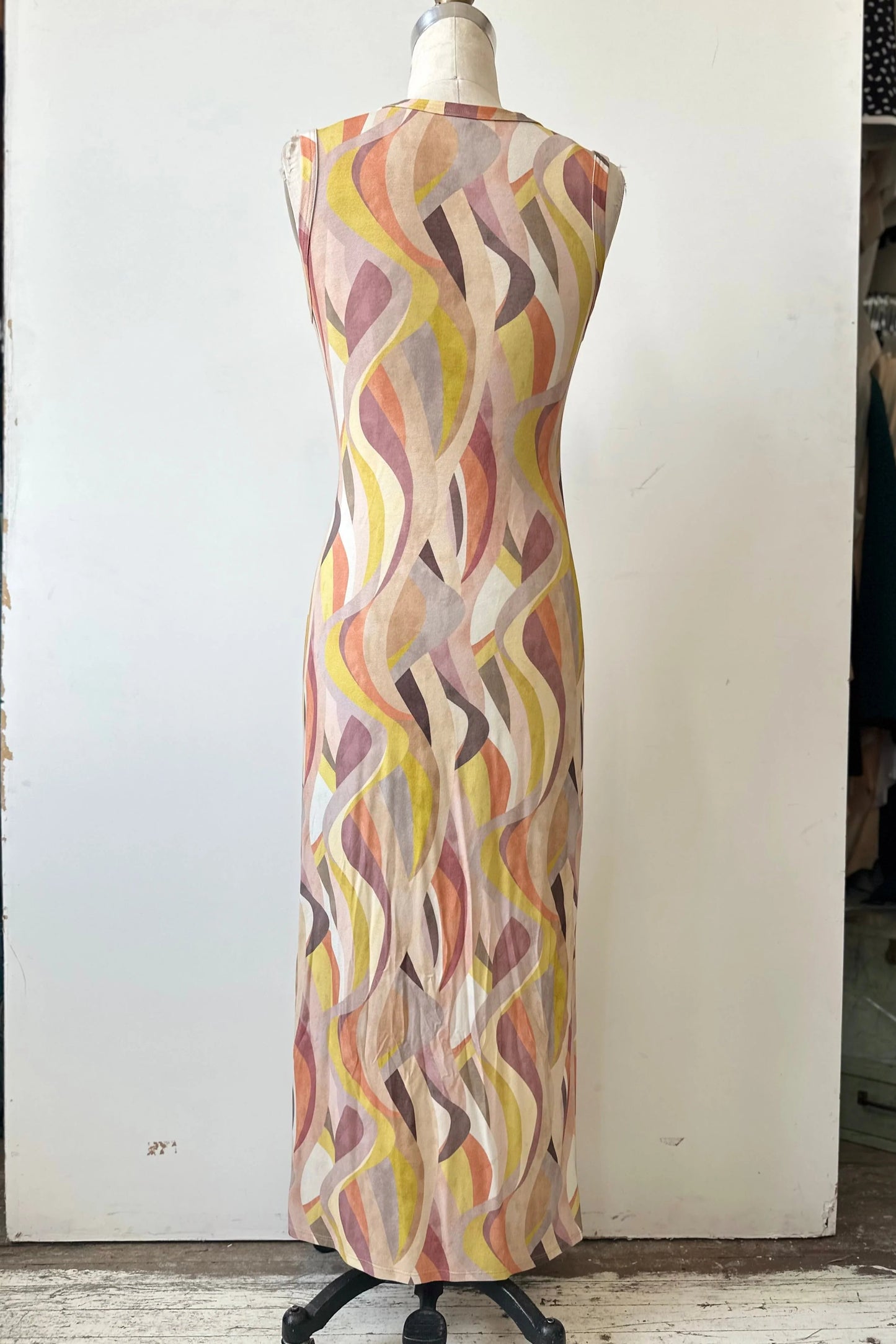A back view of the Serina Dress by Kollontai in Peach print, a long, slim cut sleeveless dress with a soft pleat at the waist, is shown on a mannequin in front of a white background. 