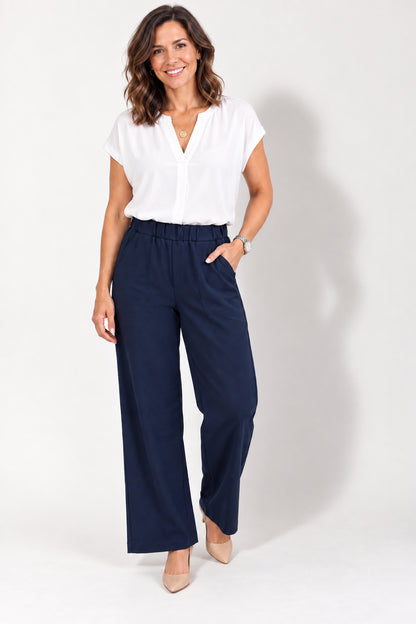 A woman wearing the Luisa Ponte Pants by Tangente in Navy, pull-on pants with an elastic waist, front pleats, and side pockets. She is wearing them with a white top and standing in front of a white studio background. 