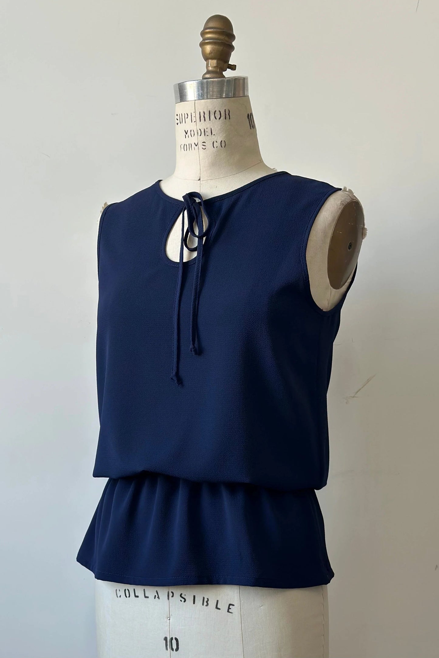 The Serena Top from Kollontai in Navy, a sleeveless top with a keyhole and tie neckline, elastic waist and flared peplum hem, is shown on a mannequin against a white background. 