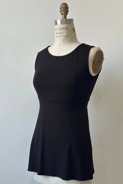 The Nira Top by Kollontai in Black, a sleeveless top with a wide neck, an empire waist, and a cut that's fitted through the bodice and flares away from the torso, is shown on a mannequin against a white background. 