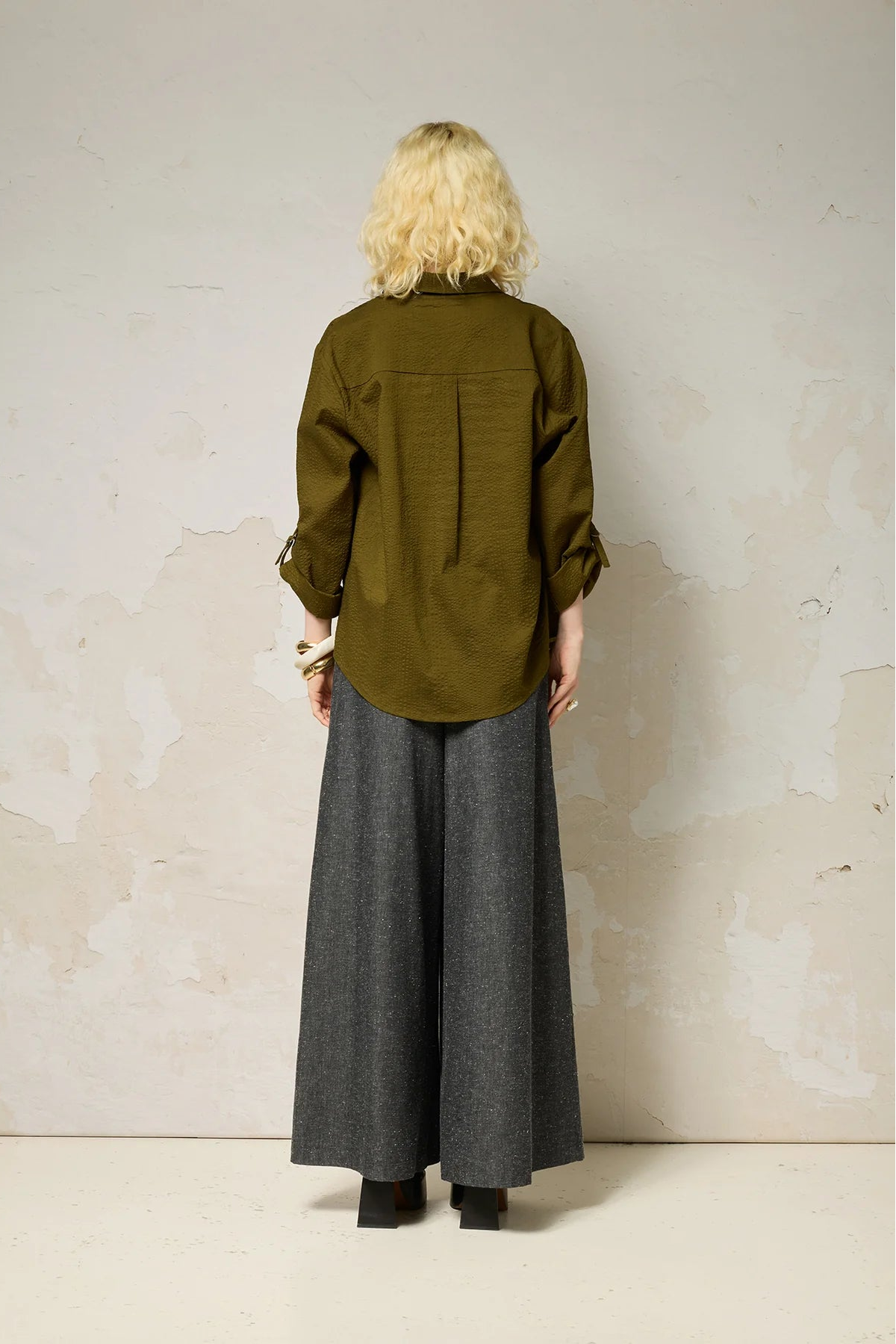 A woman wearing the Ania Shirt from Cokluch in Olive, a button front collared shirt with adjustable tab and ring details at the sleeves and a rounded hip-length hem. She is wearing it with denim pants and standing in front of a textured wall. 
