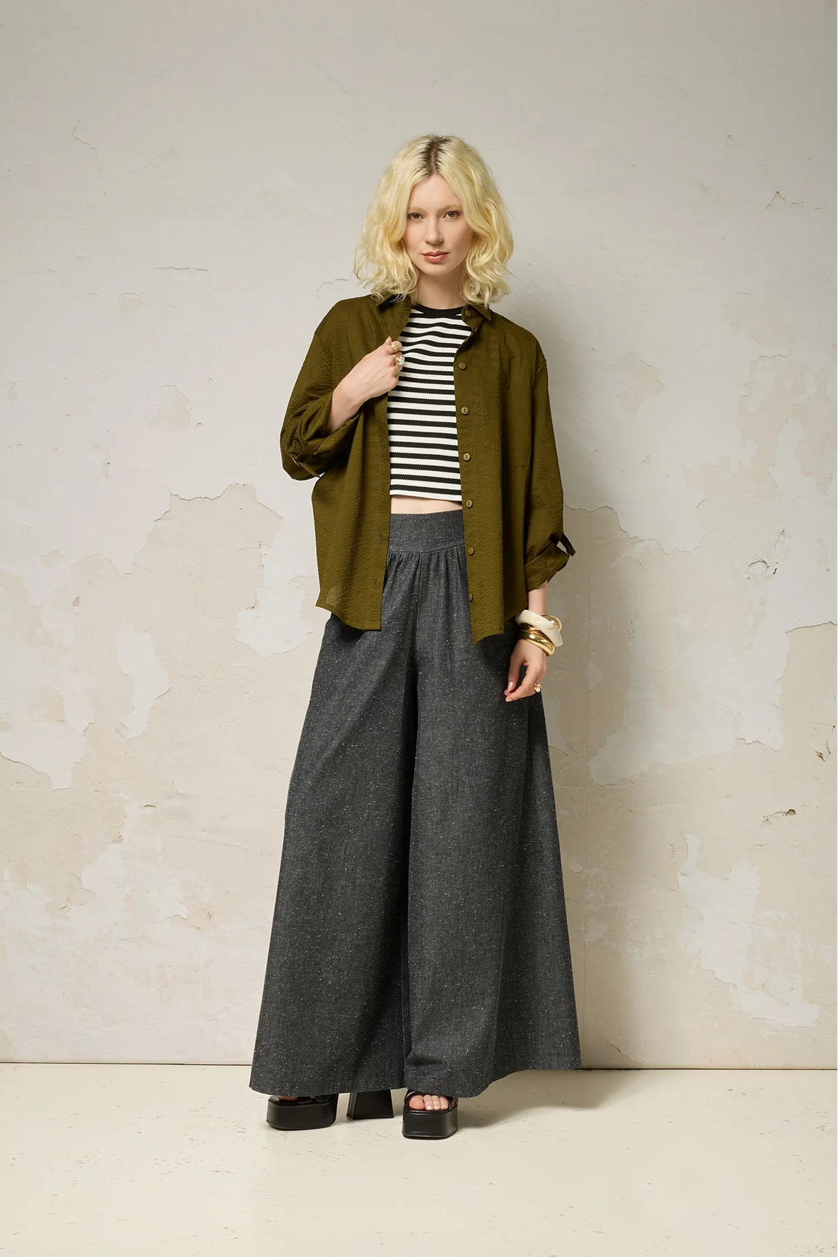 A woman wearing the Ania Shirt from Cokluch in Olive, a button front collared shirt with adjustable tab and ring details at the sleeves and a rounded hip-length hem. She is wearing it with a striped cropped top and denim pants and standing in front of a textured wall. 
