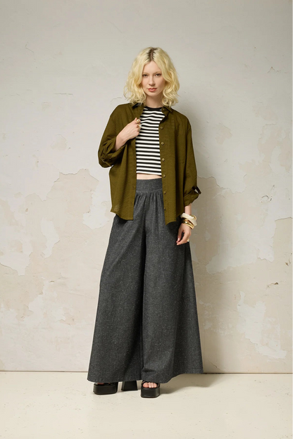 A woman wearing the Ania Shirt from Cokluch in Olive, a button front collared shirt with adjustable tab and ring details at the sleeves and a rounded hip-length hem. She is wearing it with a striped cropped top and denim pants and standing in front of a textured wall. 
