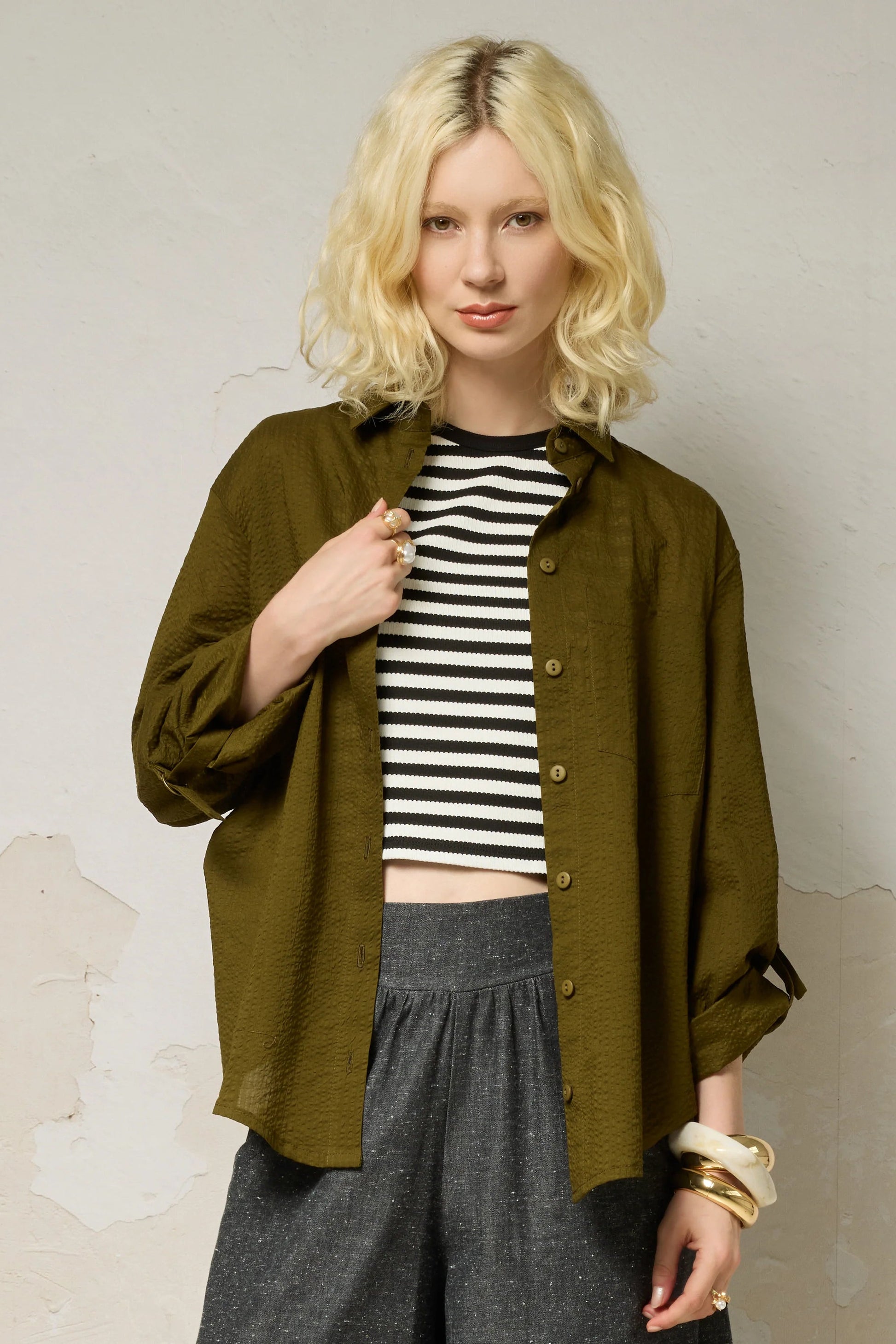 3/4 view of a woman wearing the Ania Shirt from Cokluch in Olive, a button front collared shirt with adjustable tab and ring details at the sleeves and a rounded hip-length hem. She is wearing it with a striped cropped top and denim pants and standing in front of a textured wall. 