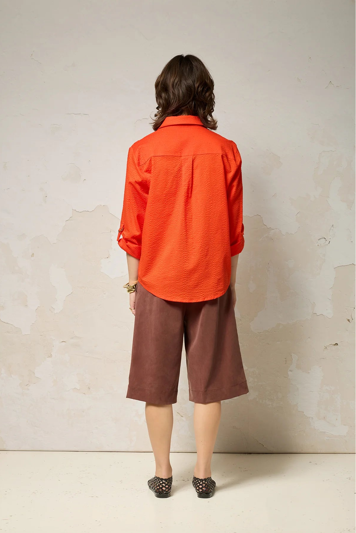 Back view of a woman wearing the Ania Shirt from Cokluch in Fiesta, a button front collared shirt with adjustable tab and ring details at the sleeves and a rounded hip-length hem. She is wearing it with brown shorts and standing against a textured wall.