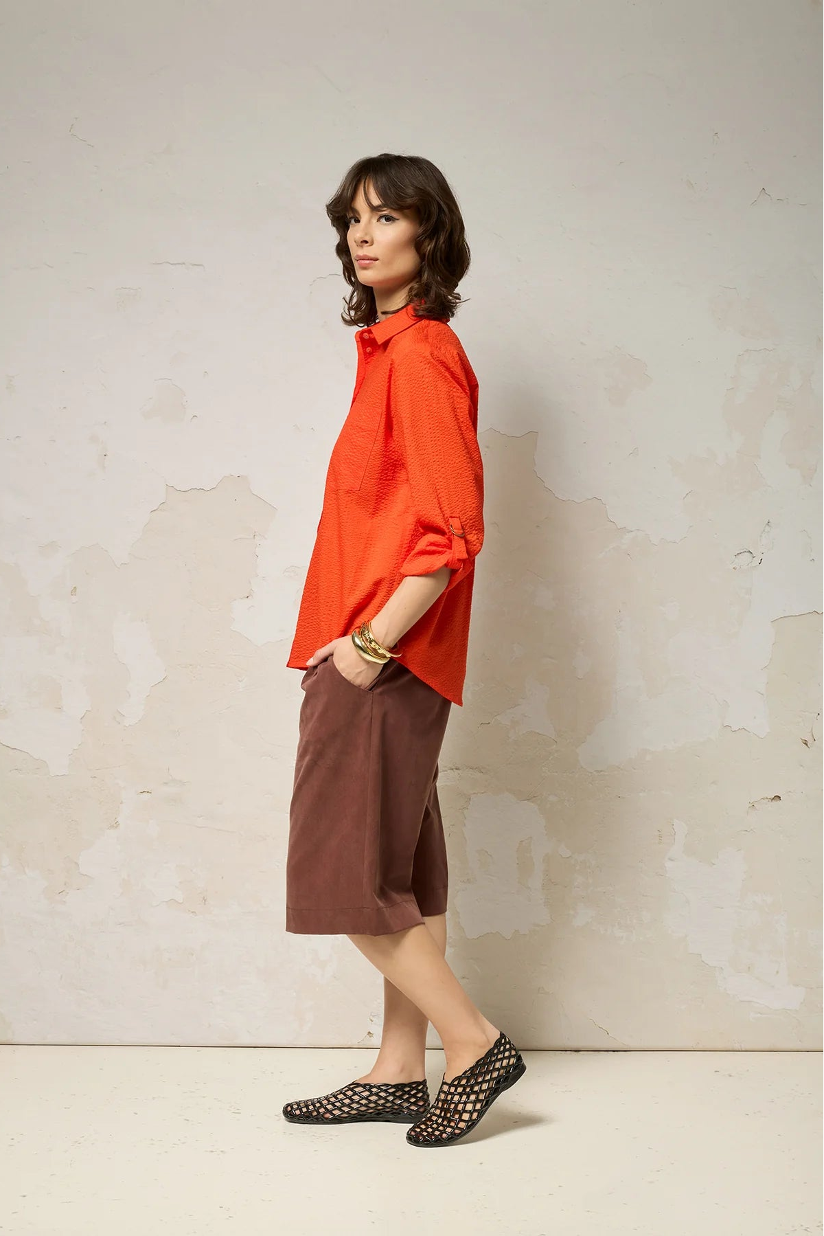 Side view of a woman wearing the Ania Shirt from Cokluch in Fiesta, a button front collared shirt with adjustable tab and ring details at the sleeves and a rounded hip-length hem. She is wearing it with brown shorts and standing against a textured wall.