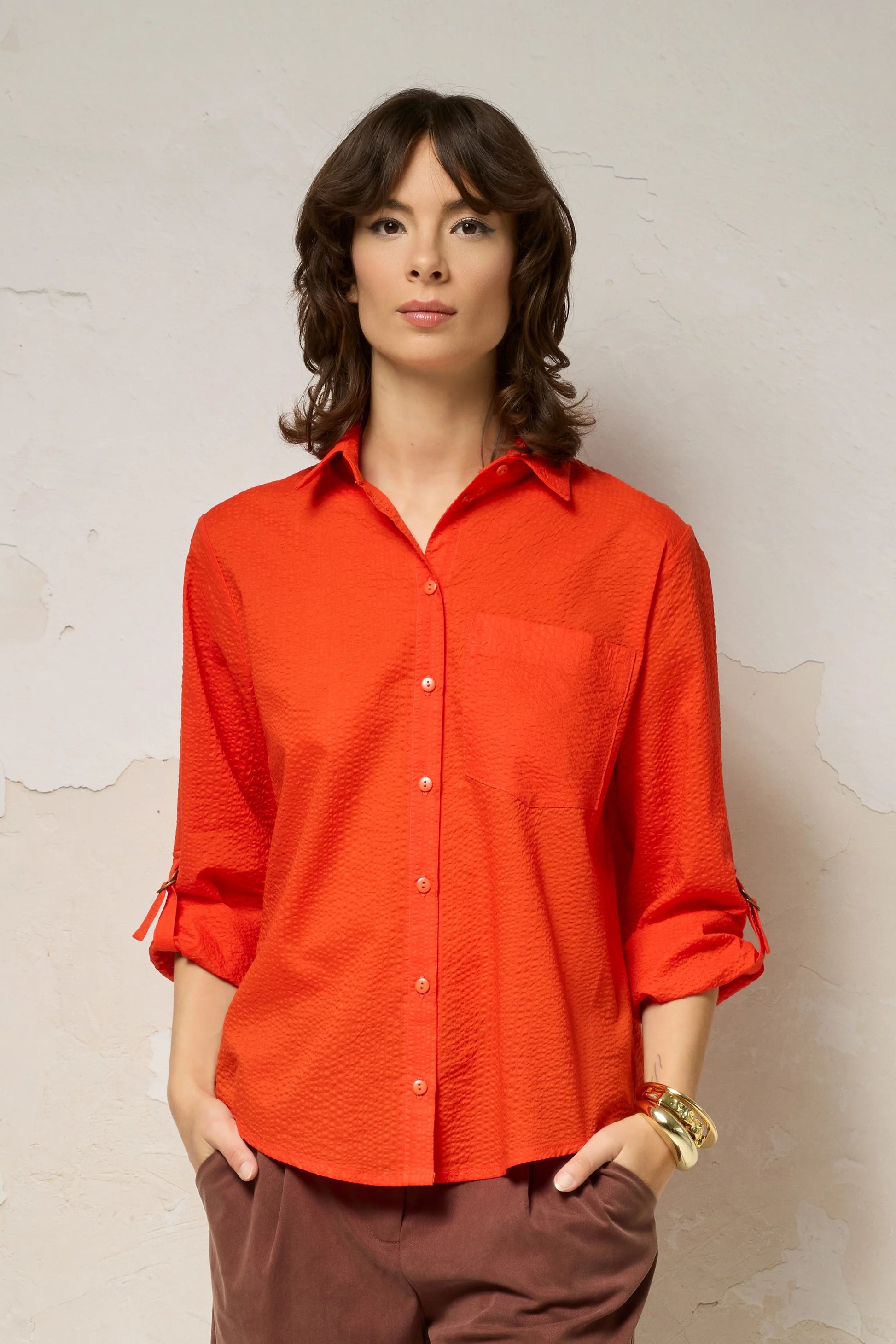 3/4 view of a woman wearing the Ania Shirt from Cokluch in Fiesta, a button front collared shirt with adjustable tab and ring details at the sleeves and a rounded hip-length hem. She is wearing it with brown shorts and standing against a textured wall.