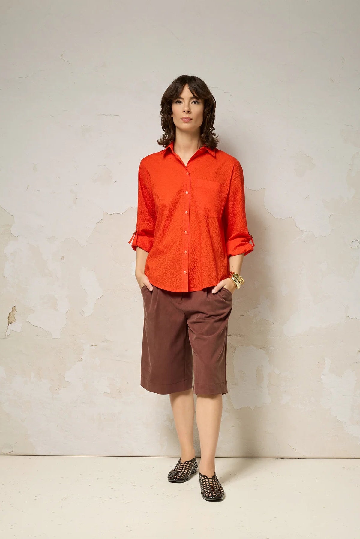 A woman wearing the Ania Shirt from Cokluch in Fiesta, a button front collared shirt with adjustable tab and ring details at the sleeves and a rounded hip-length hem. She is wearing it with brown shorts and standing against a textured wall.