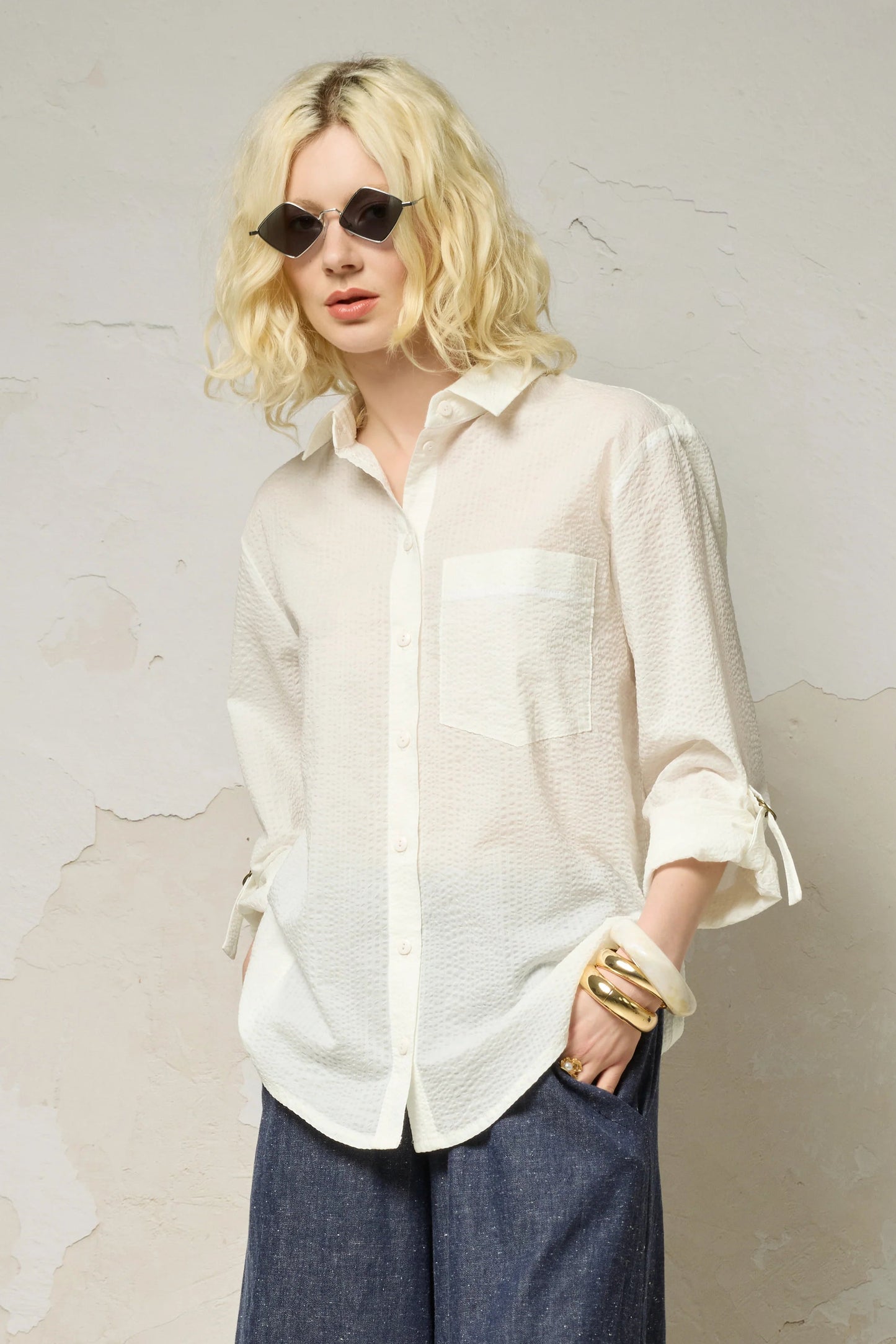 3/4 view of a woman wearing the Ania Shirt from Cokluch in Off-White, a button front collared shirt with adjustable tab and ring details at the sleeves and a rounded hip-length hem. She is wearing it with denim pants and  sunglasses and standing against a textured wall.