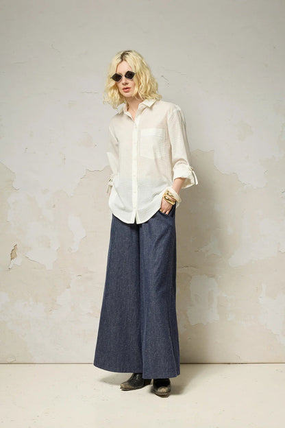 A woman wearing the Ania Shirt from Cokluch in Fiesta, a button front collared shirt with adjustable tab and ring details at the sleeves and a rounded hip-length hem. She is wearing it sunglasses and denim pants and sunglasses and standing against a textured wall.