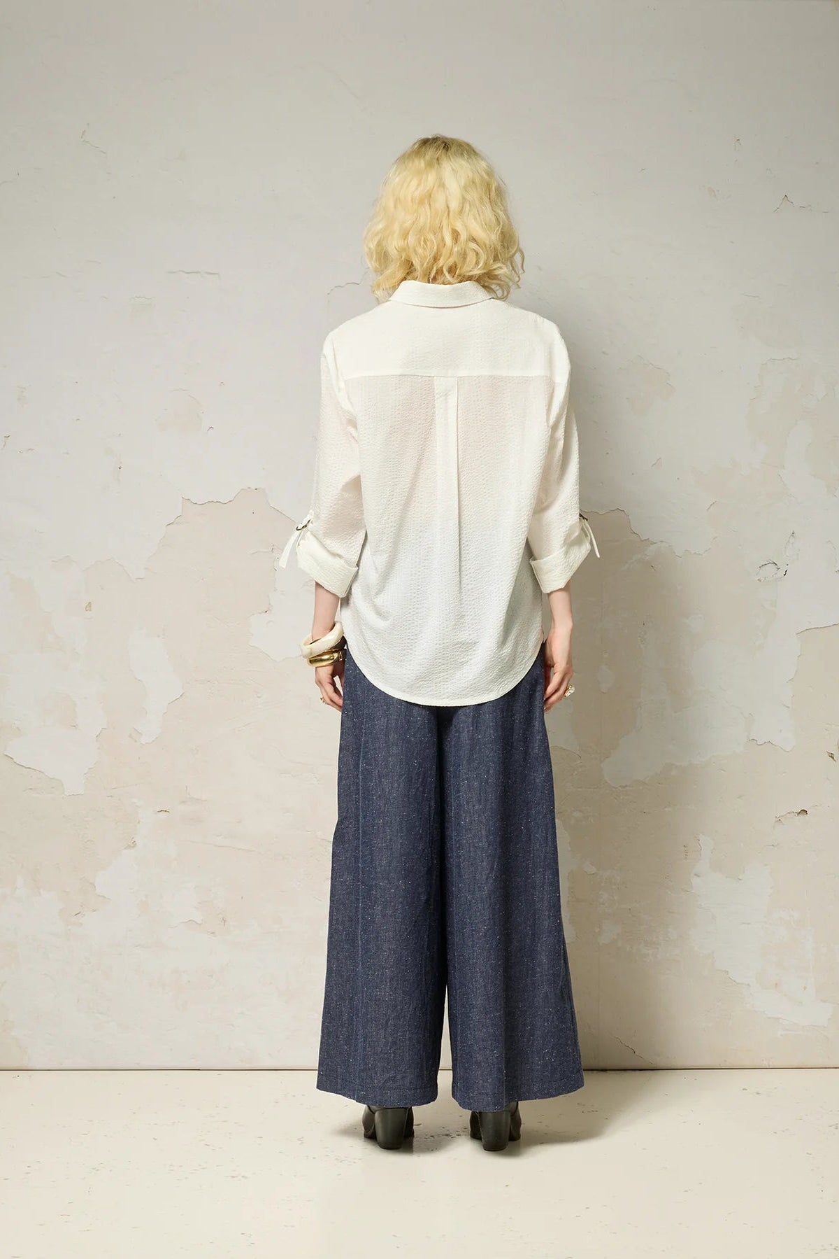 Back view of a woman wearing the Ania Shirt from Cokluch in Fiesta, a button front collared shirt with adjustable tab and ring details at the sleeves and a rounded hip-length hem. She is wearing it sunglasses and denim pants and standing against a textured wall.