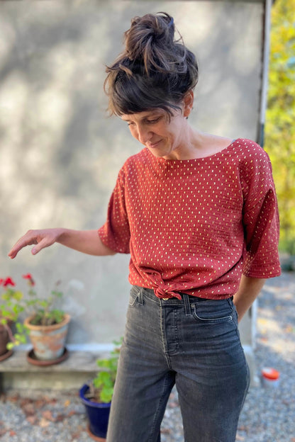 A woman wearing the Abelia Top by Kazak in Golden Tomettes, a boxy shirt with a wide neck and 3/4 kimono sleeves featuring a.red base with gold flecks. She is wearing it with grey jeans and standing outside against a grey wall. 