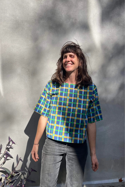 A woman wearing the Abelia Top by Kazak in Blue checks, a boxy shirt with a wide neck and 3/4 kimono sleeves. She is wearing it with grey jeans and standing outside against a grey wall. 
