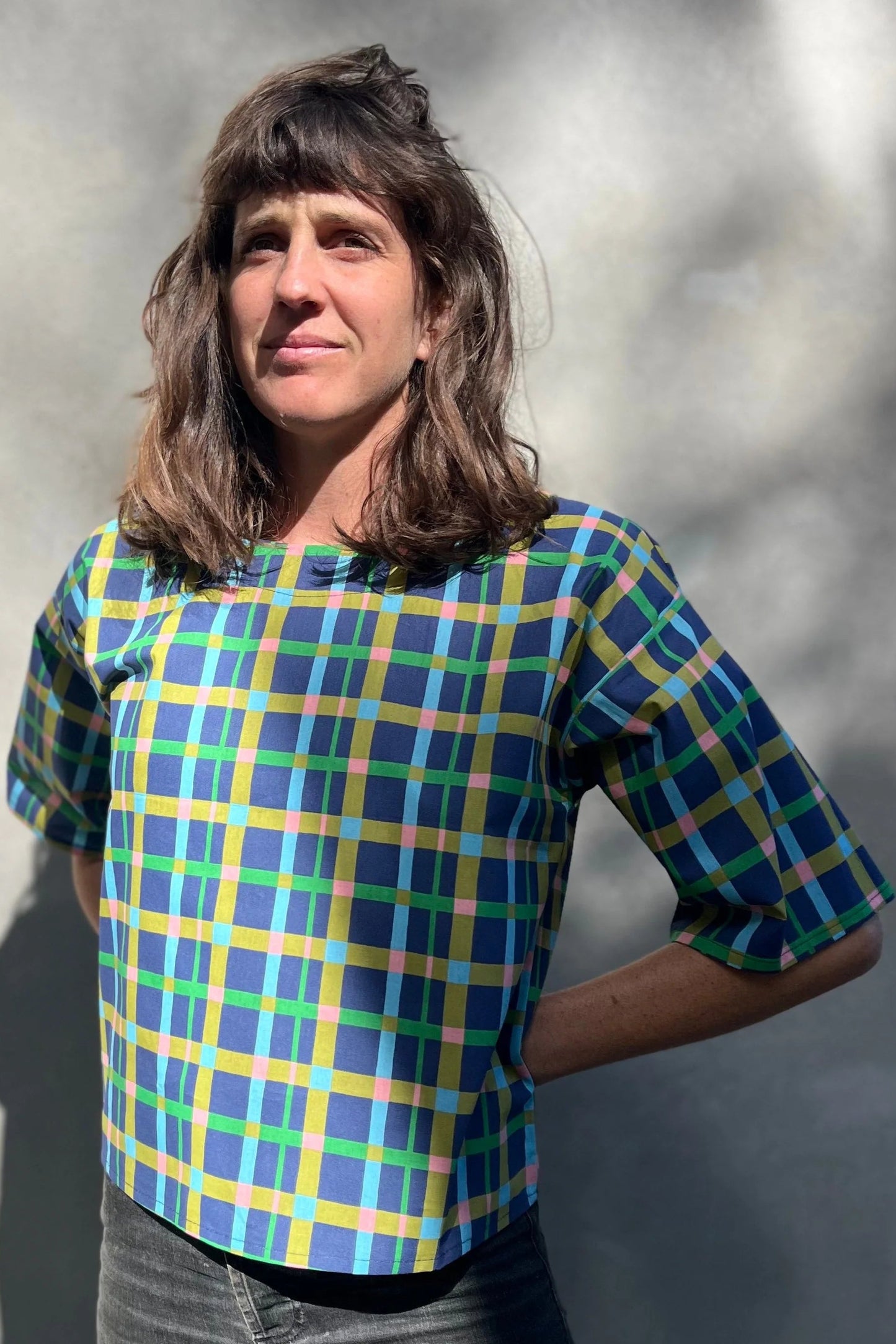 A woman wearing the Abelia Top by Kazak in Blue checks, a boxy shirt with a wide neck and 3/4 kimono sleeves. She is wearing it with grey jeans and standing outside against a grey wall. 