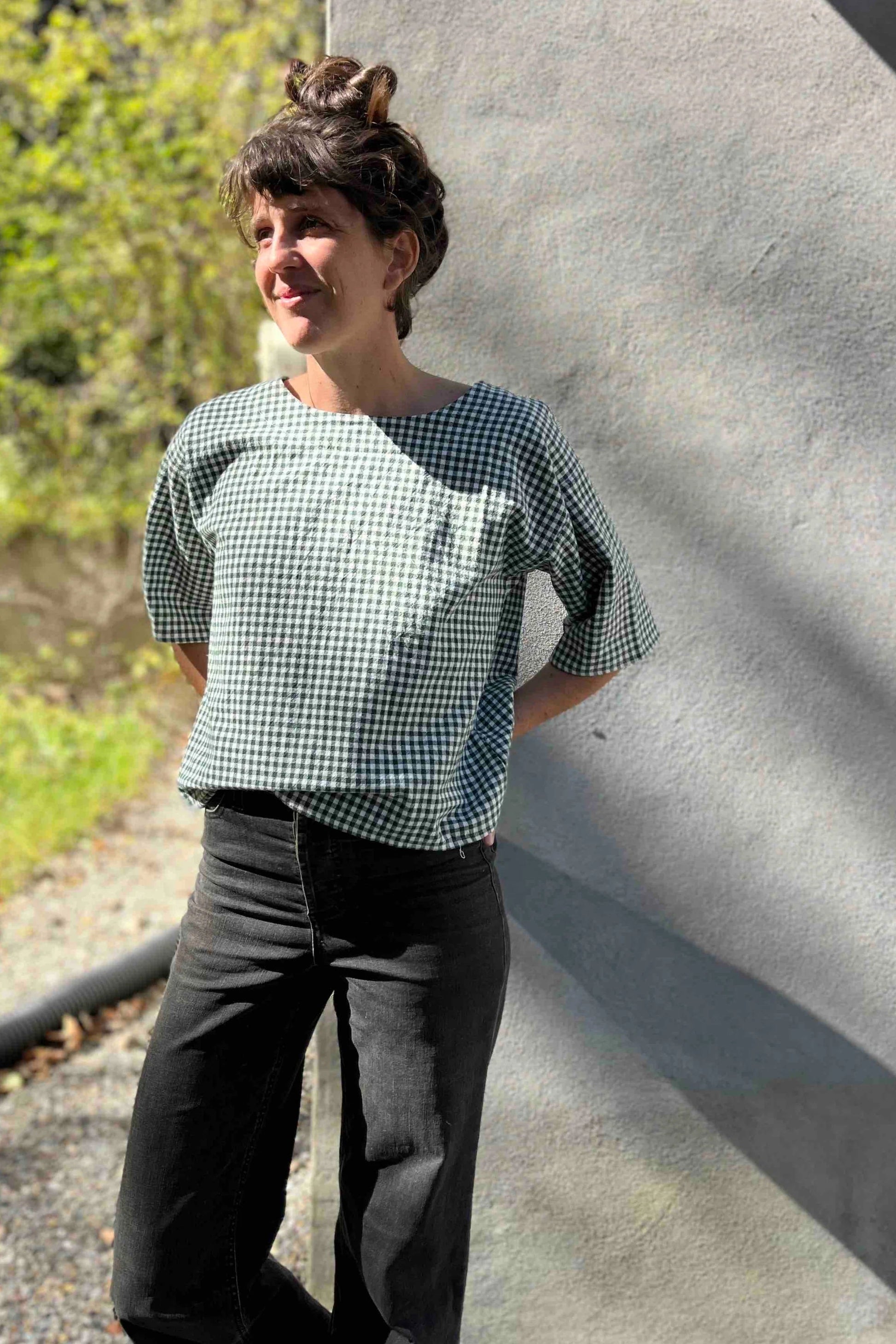 A woman wearing the Abelia Top by Kazak in Forest Vichy, a boxy shirt with a wide neck and 3/4 kimono sleeves. She is wearing it with grey jeans and standing outside.