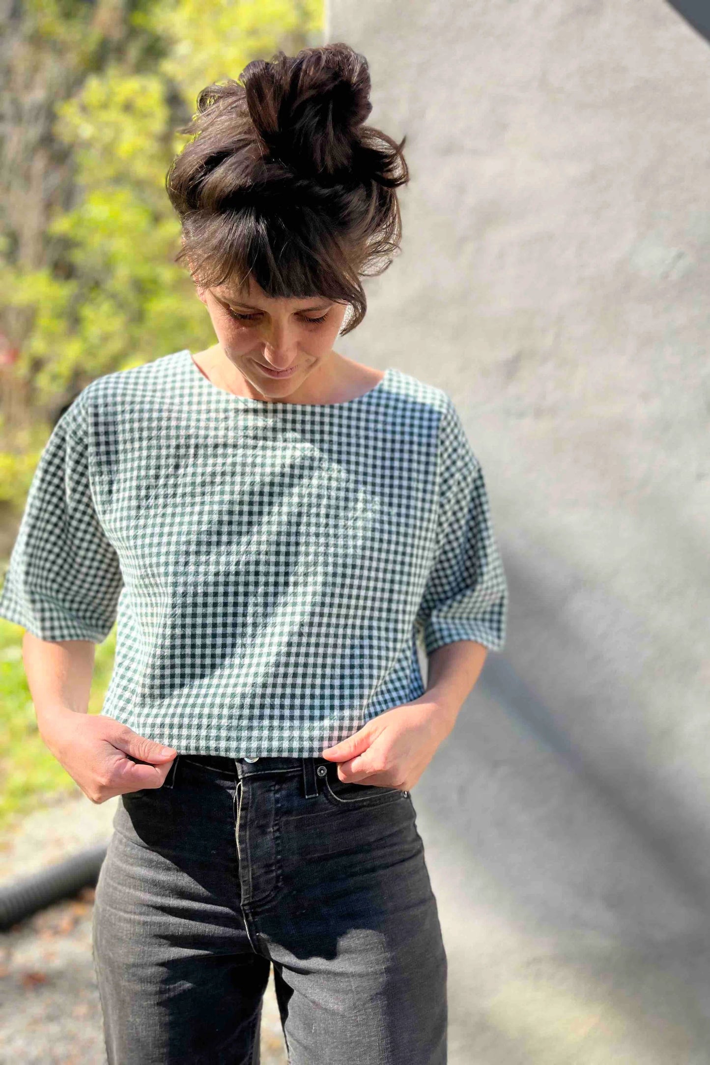 A woman wearing the Abelia Top by Kazak in Forest Vichy, a boxy shirt with a wide neck and 3/4 kimono sleeves. She is wearing it with grey jeans and standing outside against a grey wall. 