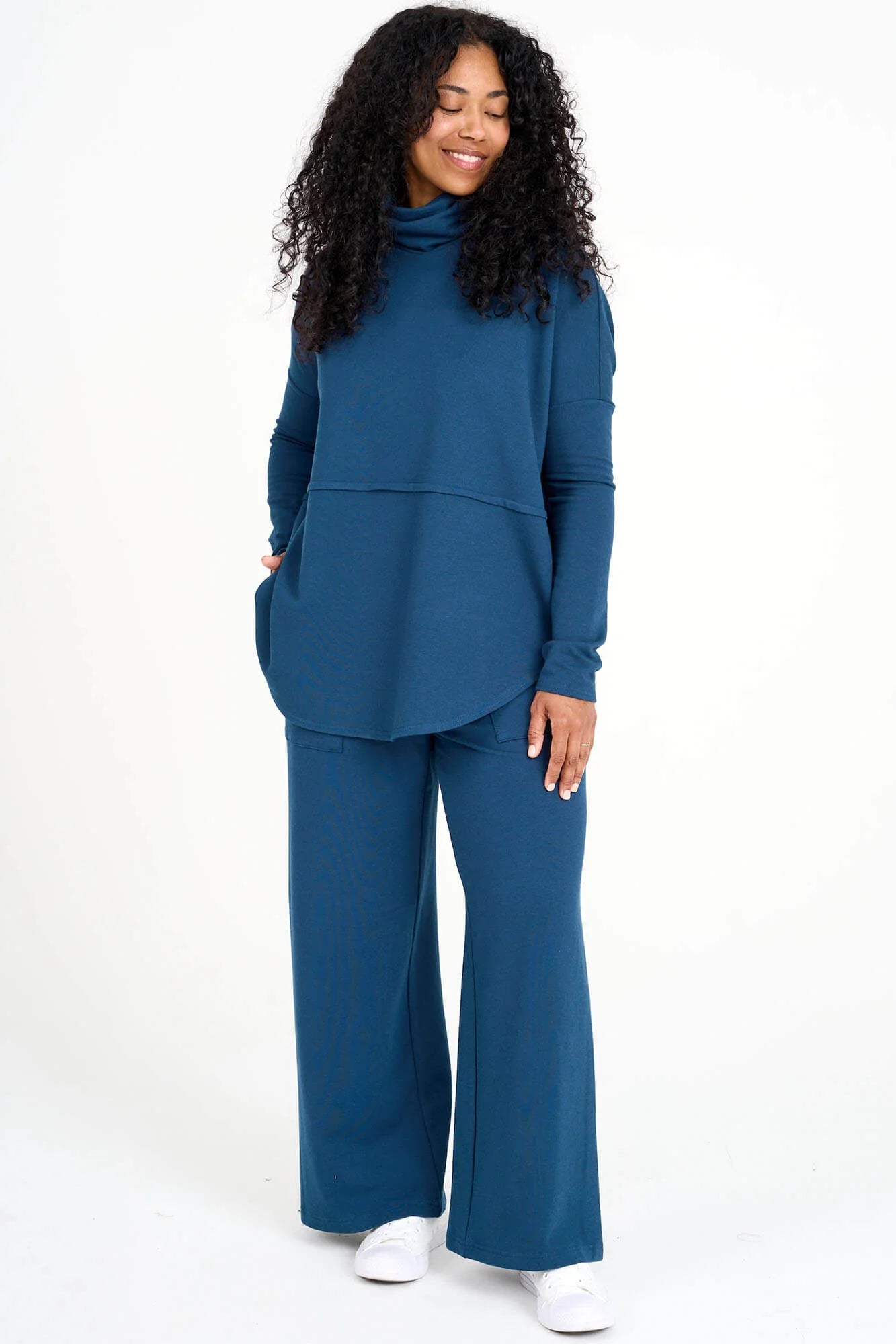 A woman wearing the Abigail Turtleneck by Advika in Bluebell, a loose fitting turtleneck top with dropped sleeves, a seam across the middle, and a rounded hem. She is wearing it with matching pants and standing in front of a white background. 