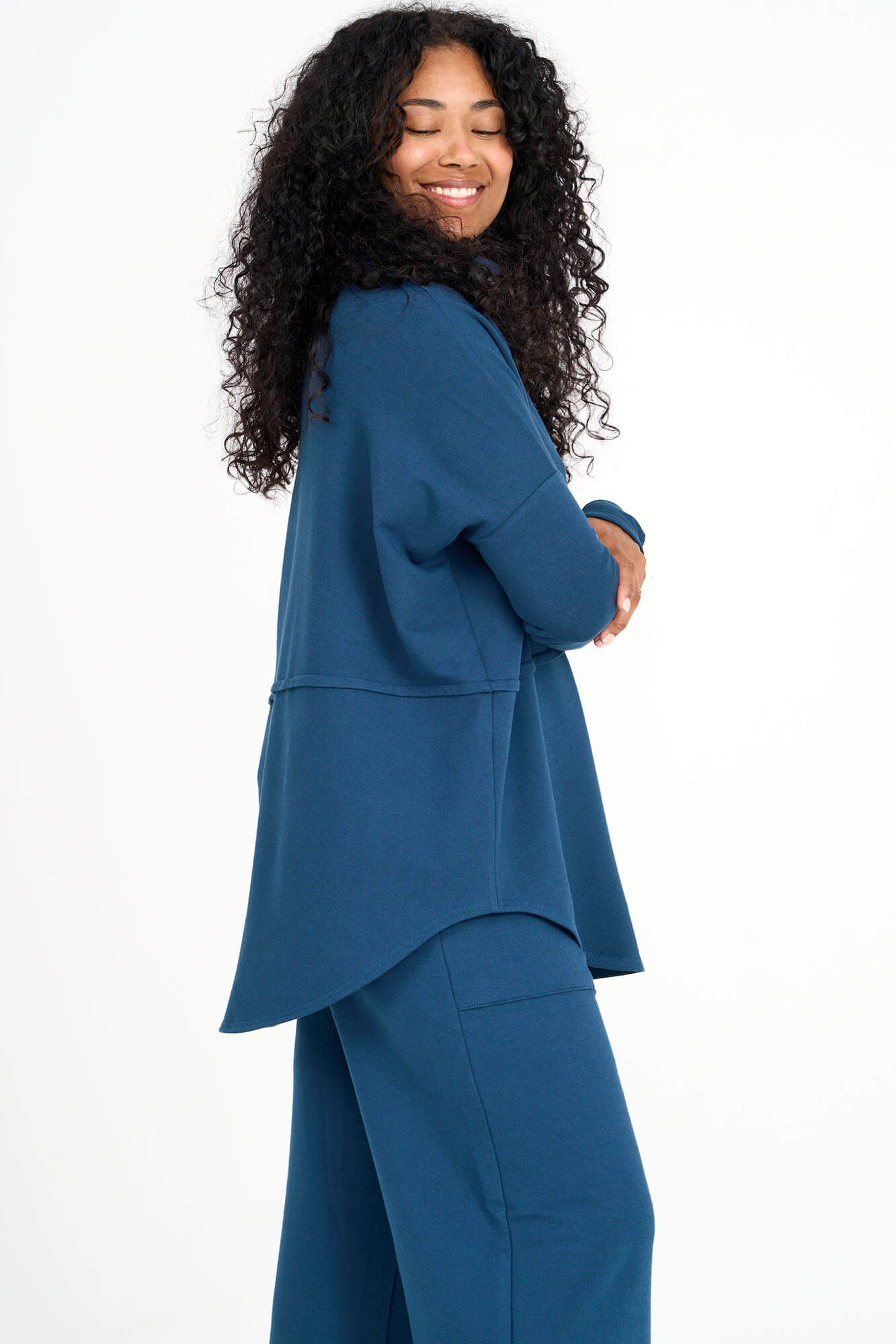 A side view of a woman wearing the Abigail Turtleneck by Advika in Bluebell, a loose fitting turtleneck top with dropped sleeves, a seam across the middle, and a rounded hem. She is wearing it with matching pants and standing in front of a white background. 