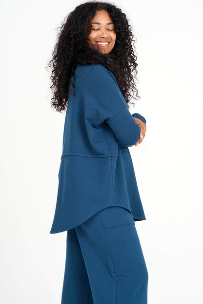 A side view of a woman wearing the Abigail Turtleneck by Advika in Bluebell, a loose fitting turtleneck top with dropped sleeves, a seam across the middle, and a rounded hem. She is wearing it with matching pants and standing in front of a white background. 