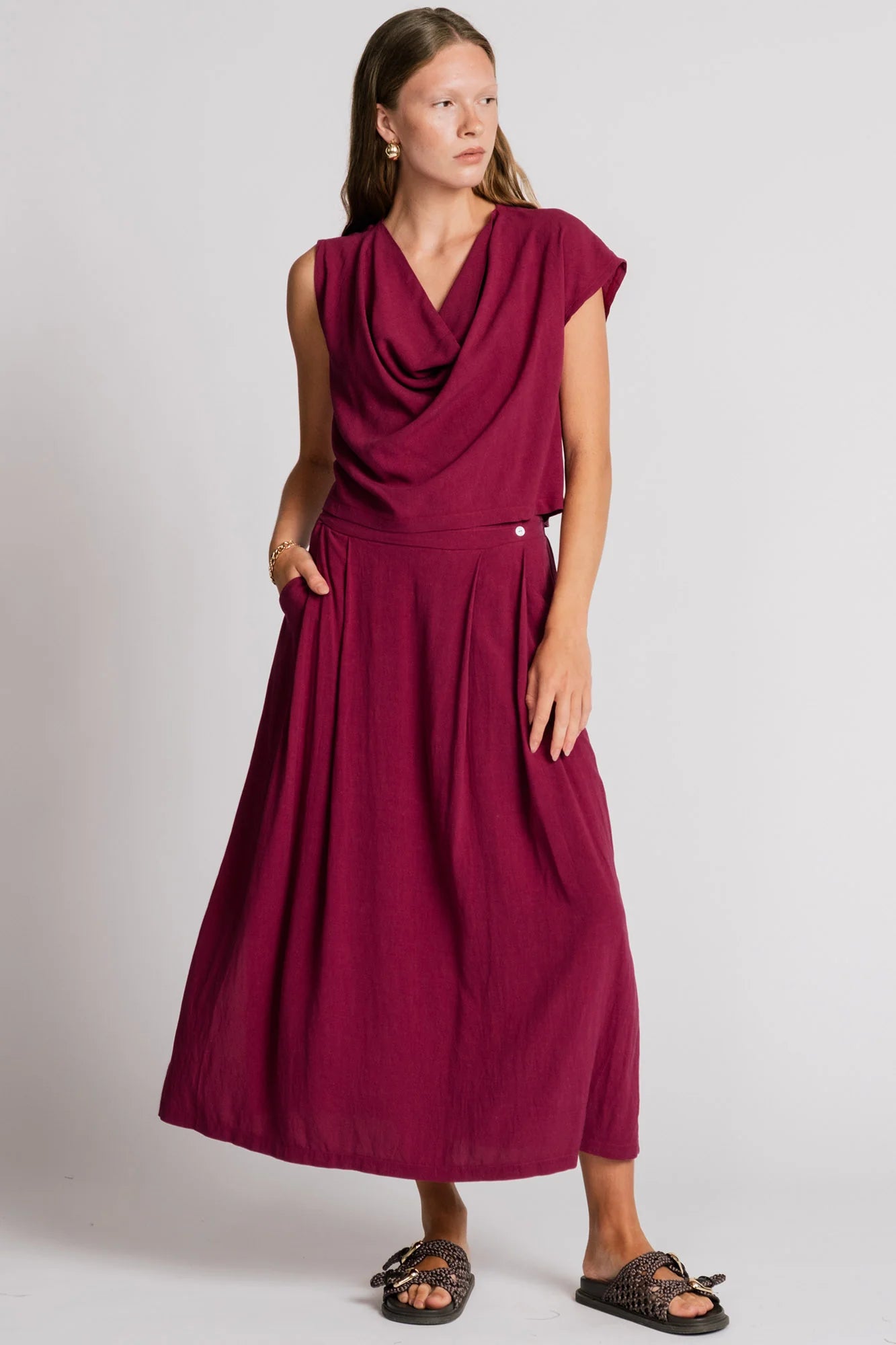 A woman wearing the Barbet Skirt by Allison Wonderland in Pink, a maxi dress with side slits, front pleats, elastic at the back waist and a side button detail at the front. She is wearing it with a matching tank and standing in front of a white background. 