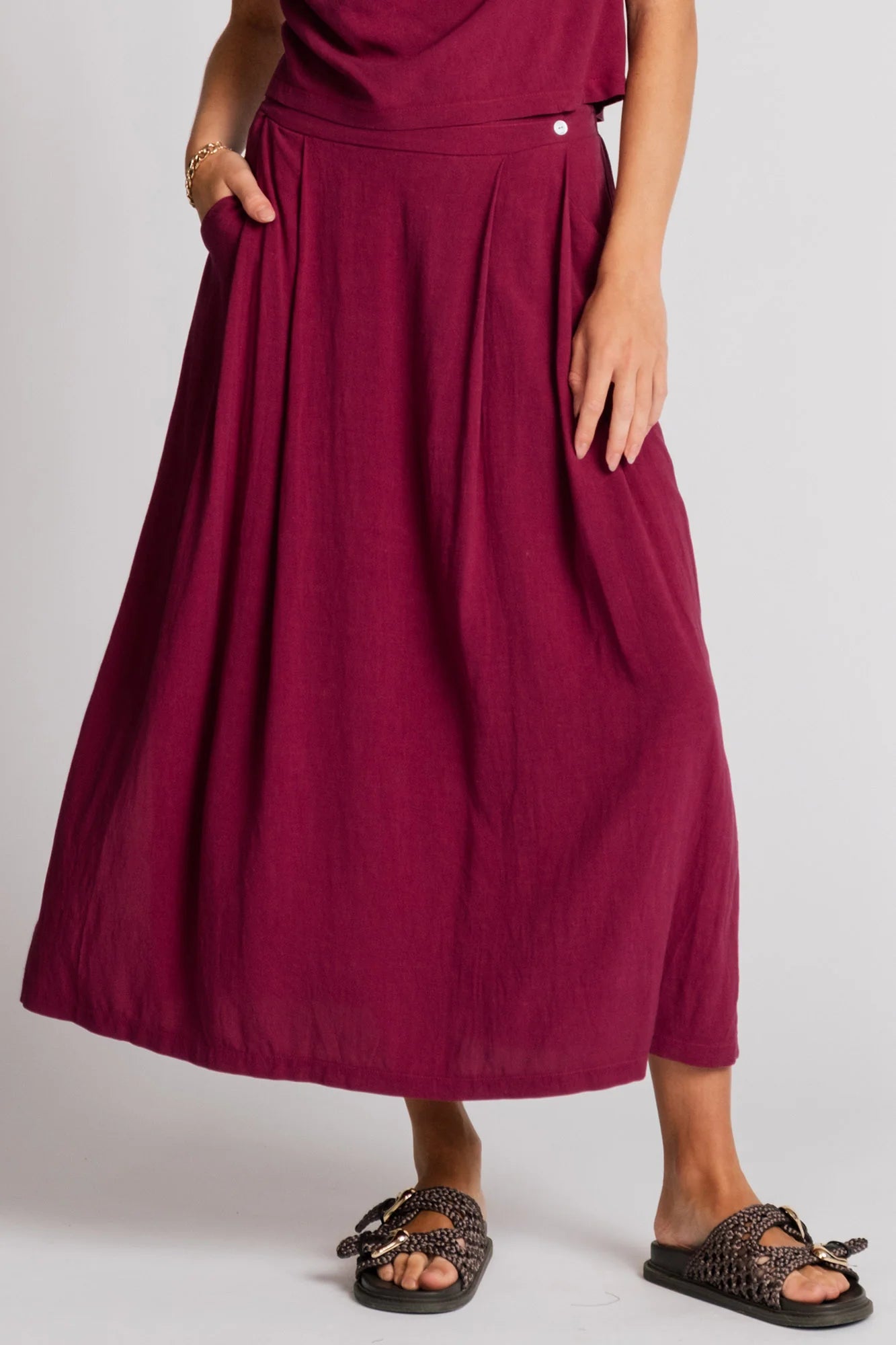 Waist down view of a woman wearing the Barbet Skirt by Allison Wonderland in Pink, a maxi dress with side slits, front pleats, elastic at the back waist and a side button detail at the front. She is wearing it with a matching tank and standing in front of a white background. 