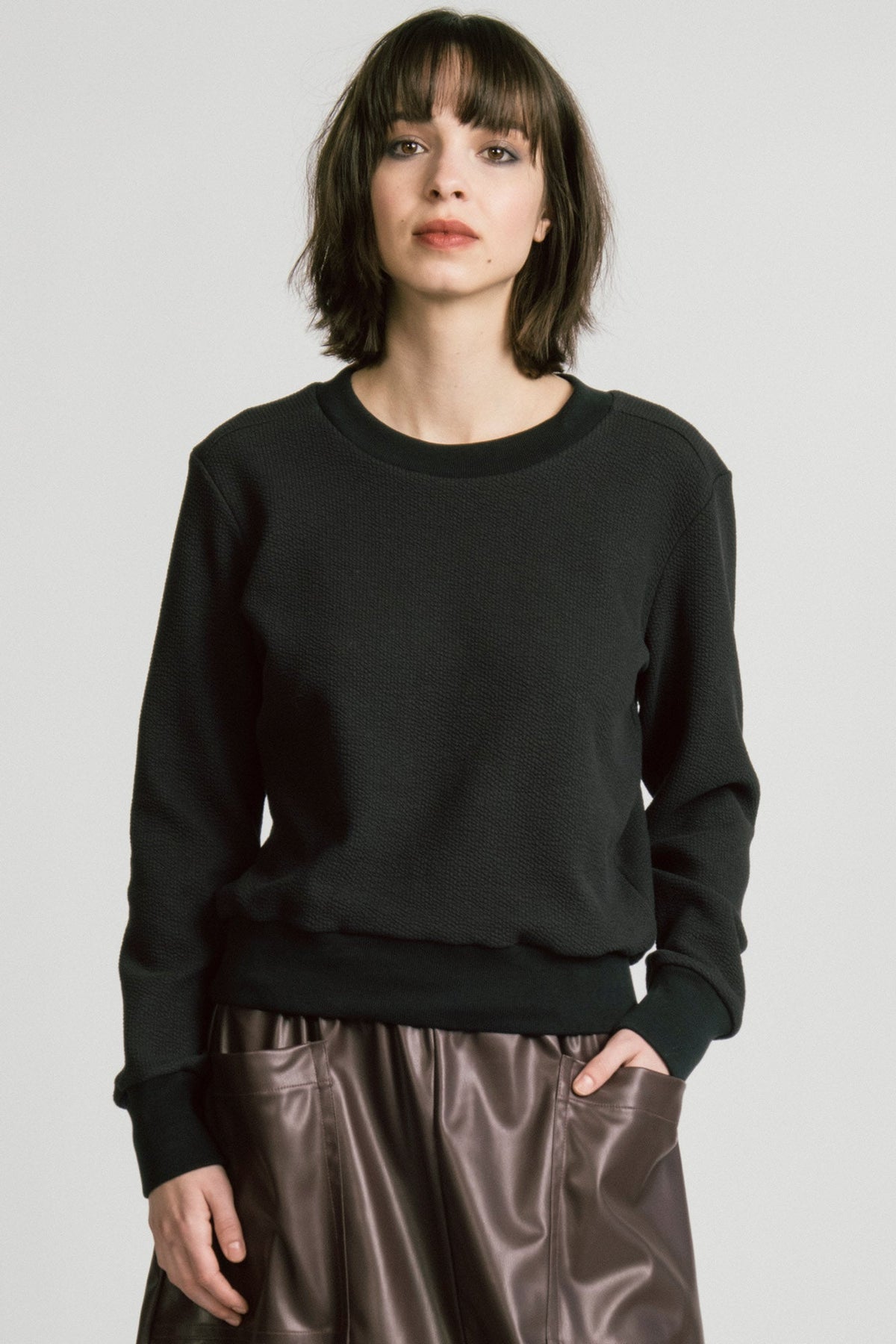 A woman wearing the Crux Sweatshirt by Allison Wonderland in Black Texture. she is wearing it with a brown pleather skirt and standing in front of a white background. 