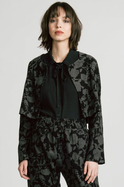 A woman wearing the Libra Bolero by Allison Wonderland in Floral, a dark printed corduroy cropped jacket with a single button closure. She is wearing it over a black top with matching pants. 