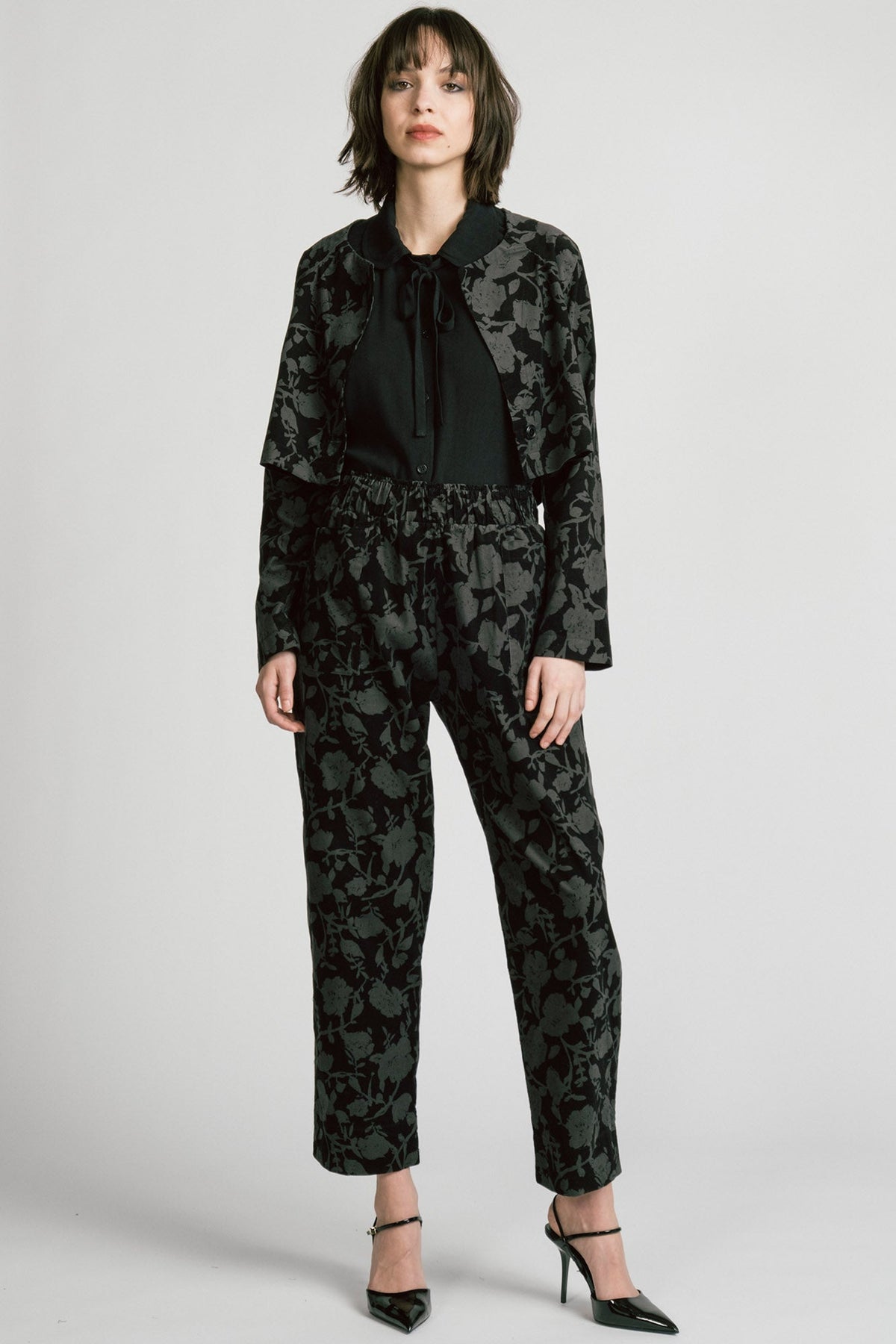 A woman wearing the Libra Bolero by Allison Wonderland in Floral, a dark printed corduroy cropped jacket with a single button closure. She is wearing it over a black top with matching pants. 