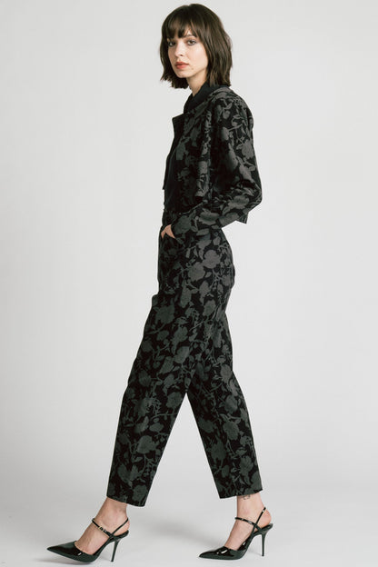 Side view of a woman wearing the Libra Bolero by Allison Wonderland in Floral, a dark printed corduroy cropped jacket with a single button closure. She is wearing it over a black top with matching pants. 