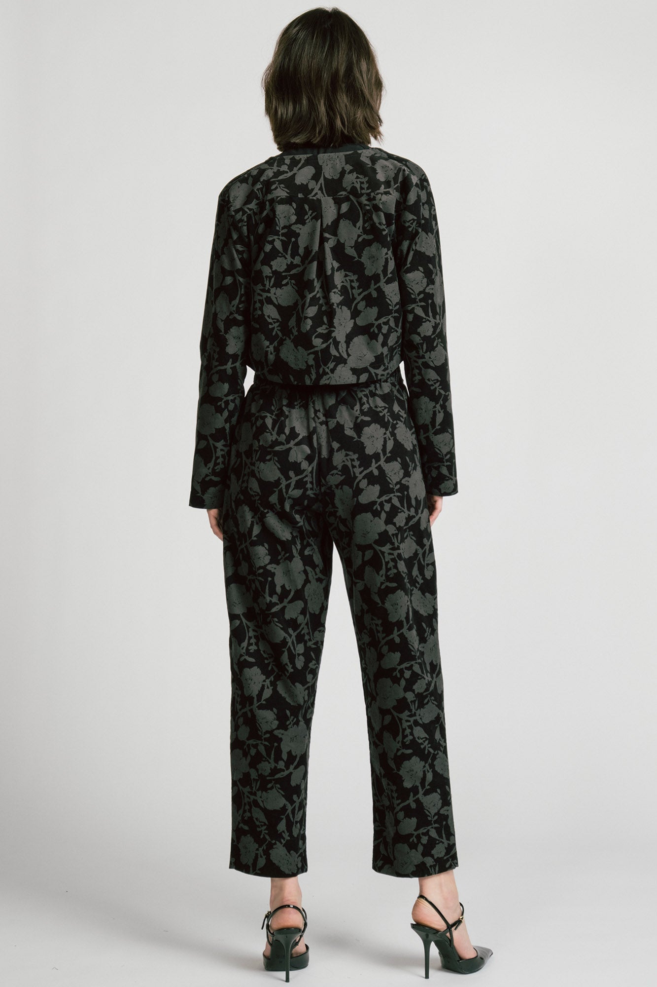 Back view of a woman wearing the Libra Bolero by Allison Wonderland in Floral, a dark printed corduroy cropped jacket with a single button closure. She is wearing it over a black top with matching pants. 
