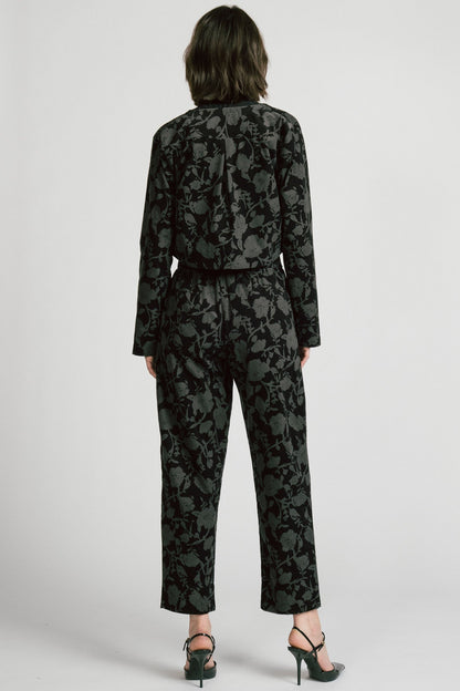 Back view of a woman wearing the Libra Bolero by Allison Wonderland in Floral, a dark printed corduroy cropped jacket with a single button closure. She is wearing it over a black top with matching pants. 