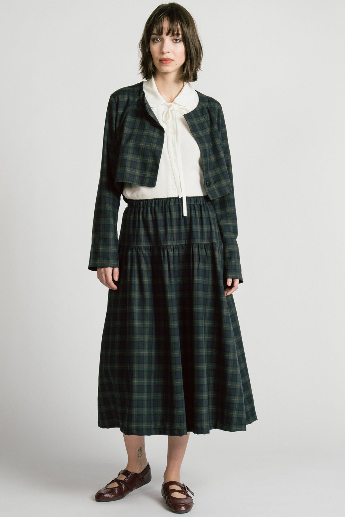 A woman wearing the Libra Bolero by Allison Wonderland in Plaid, a green plaid cropped jacket with a single button closure. She is wearing it over a white top with a matching skirt. 