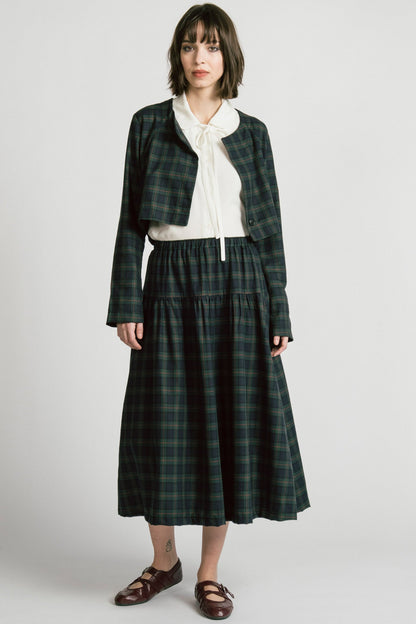 A woman wearing the Libra Bolero by Allison Wonderland in Plaid, a green plaid cropped jacket with a single button closure. She is wearing it over a white top with a matching skirt. 