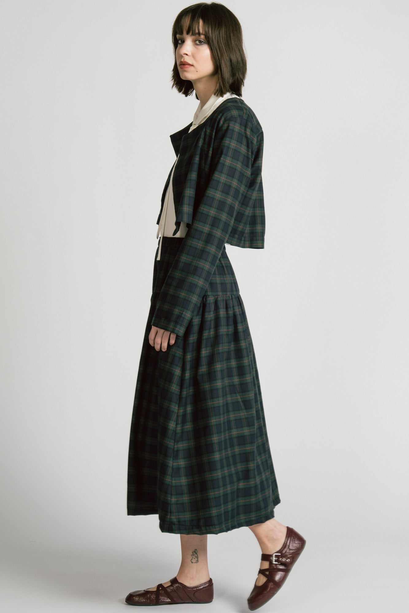 Side view of a woman wearing the Libra Bolero by Allison Wonderland in Plaid, a green plaid cropped jacket with a single button closure. She is wearing it over a white top with a matching skirt. 