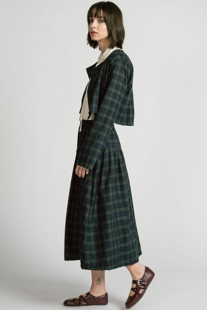 Side view of a woman wearing the Libra Bolero by Allison Wonderland in Plaid, a green plaid cropped jacket with a single button closure. She is wearing it over a white top with a matching skirt. 