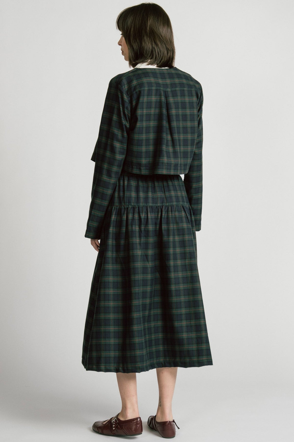 Back view of a woman wearing the Libra Bolero by Allison Wonderland in Plaid, a green plaid cropped jacket with a single button closure. She is wearing it over a white top with a matching skirt. 