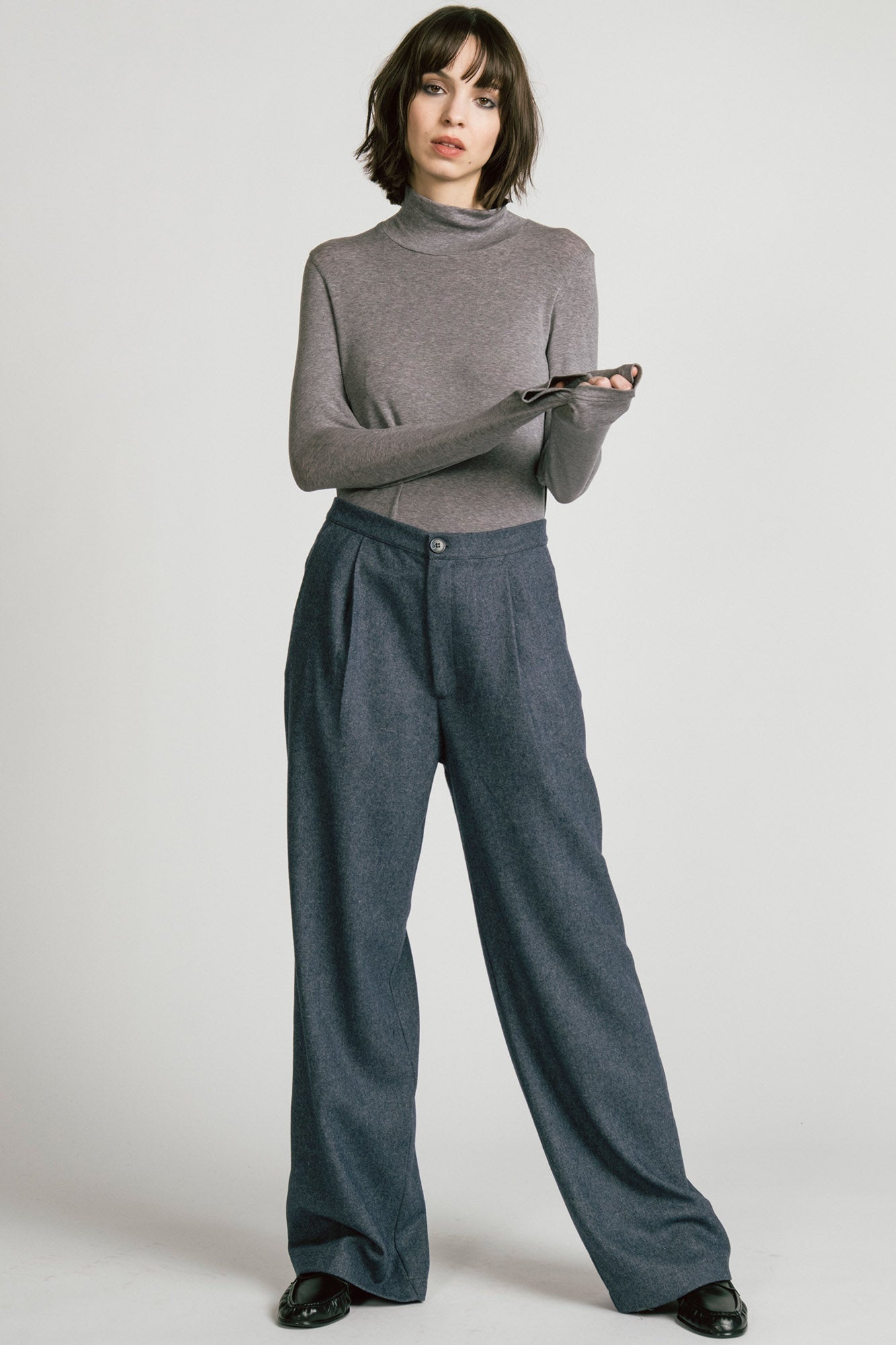 A woman wearing the Octans Turtleneck by Allison Wonderland in Heather Grey, with dark grey pleated trousers, standing against a white studio background.