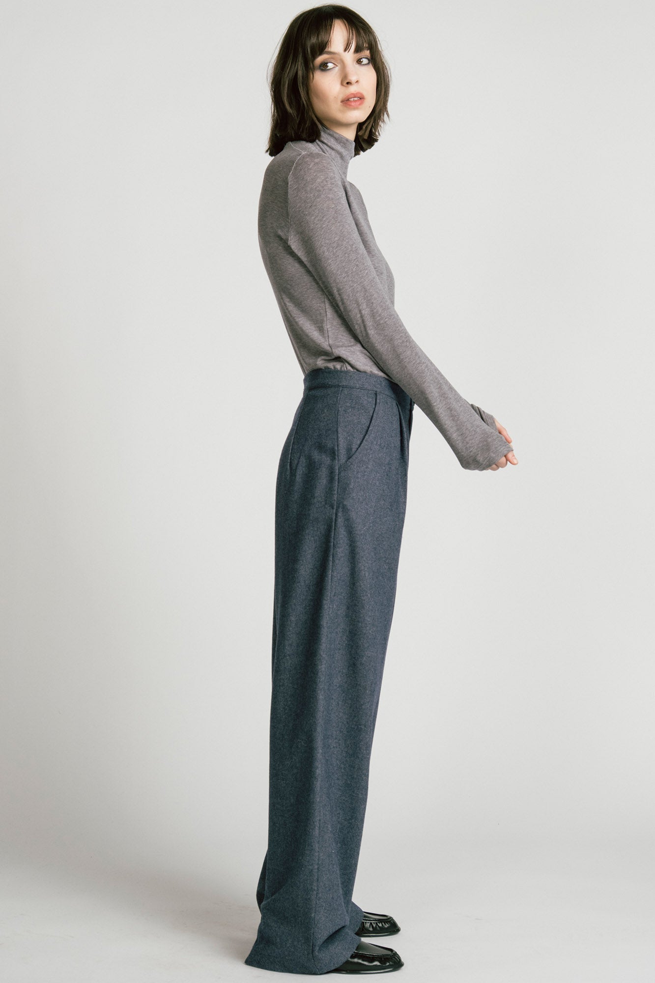 Side view of a woman wearing the Octans Turtleneck by Allison Wonderland in Heather Grey, with dark grey pleated trousers, standing against a white studio background.