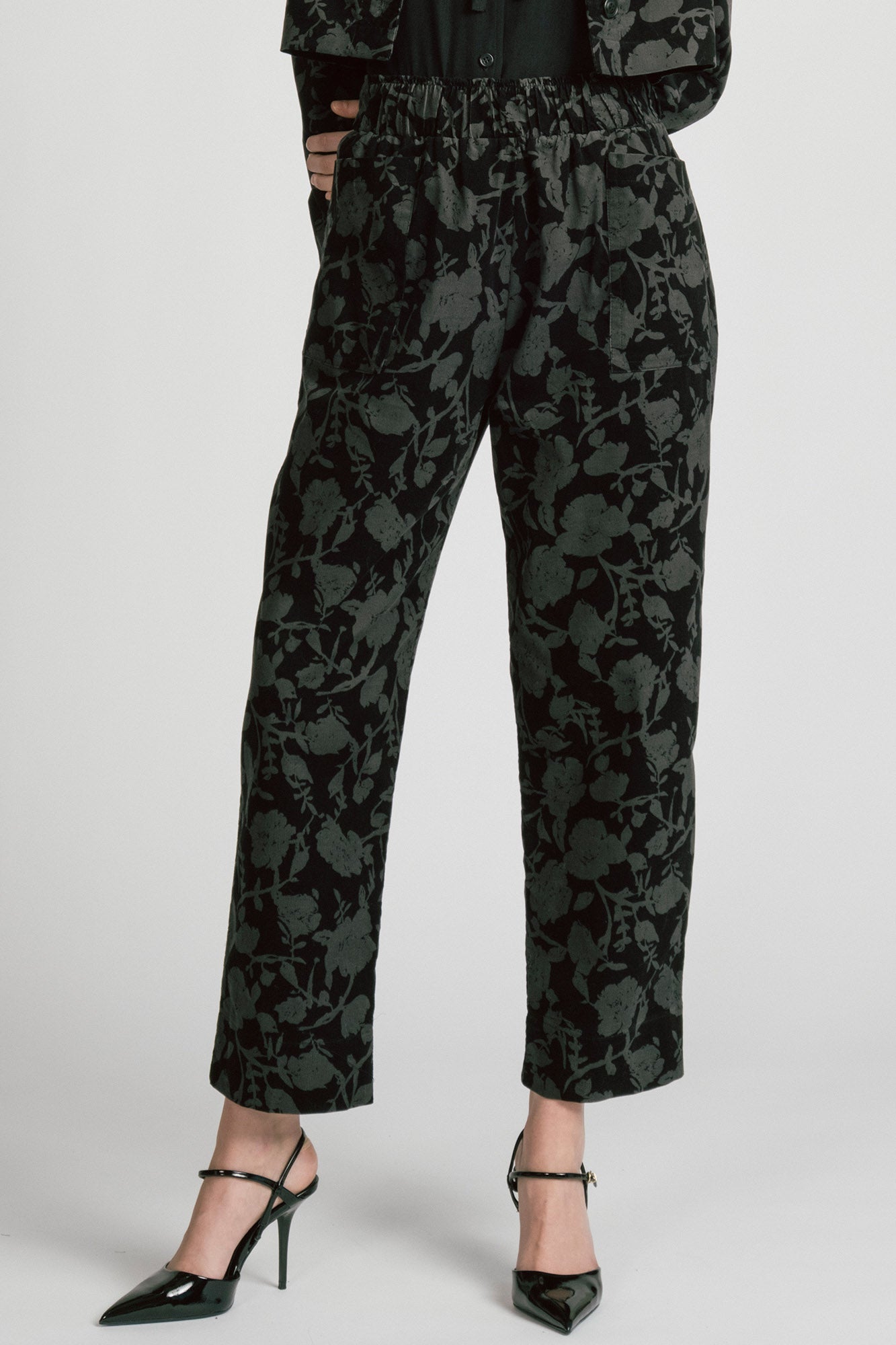 Waist down view of a woman wearing the Orion Pants by Allison Wonderland, relaxed fitting pants with an elastic waist and pockets in a dark Floral print on fine wale corduroy. She is wearing them with the matching Libra Bolero and standing in front of a white background. 
