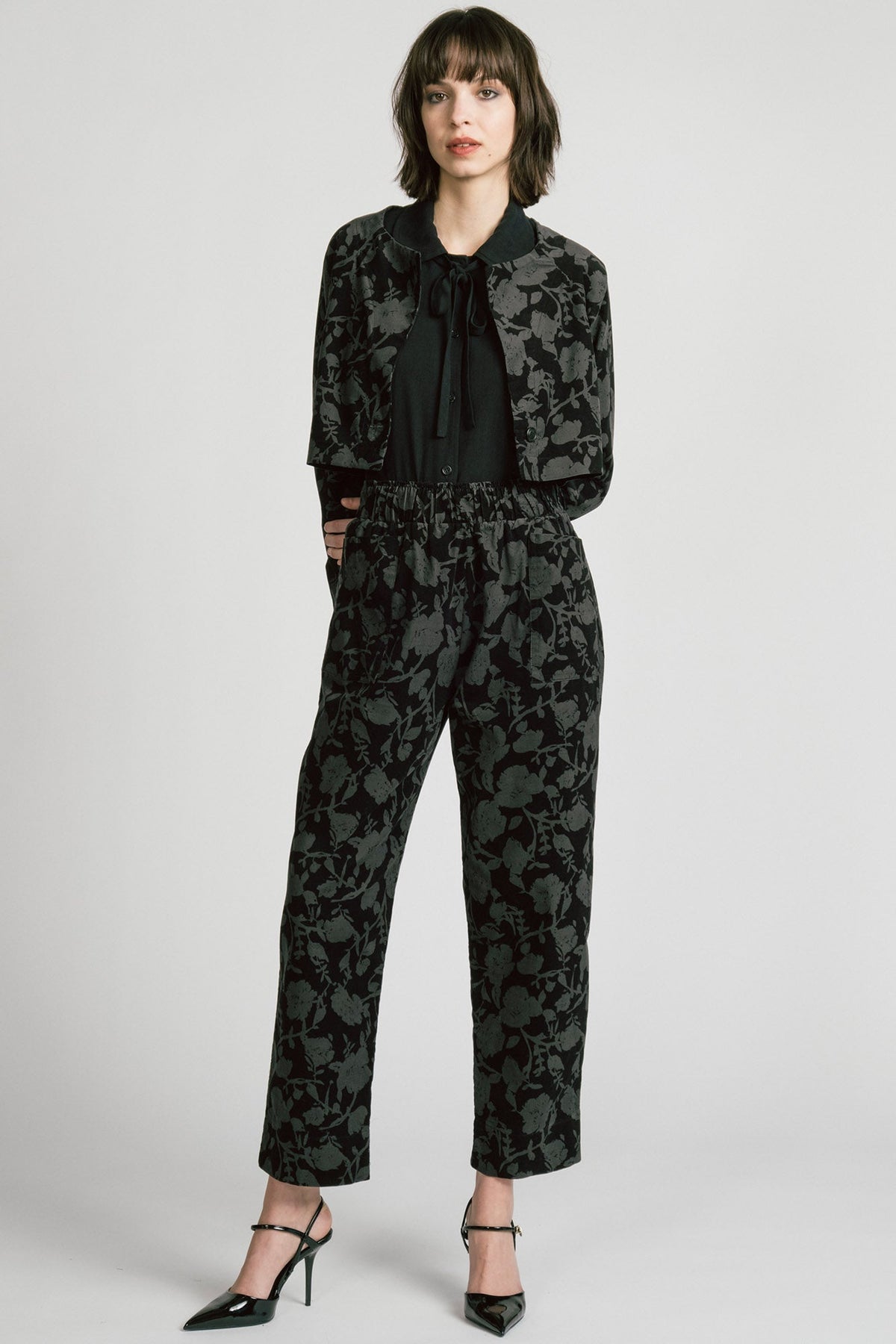 A woman wearing the Orion Pants by Allison Wonderland, relaxed fitting pants with an elastic waist and pockets in a dark Floral print on fine wale corduroy. She is wearing them with the matching Libra Bolero and standing in front of a white background. 