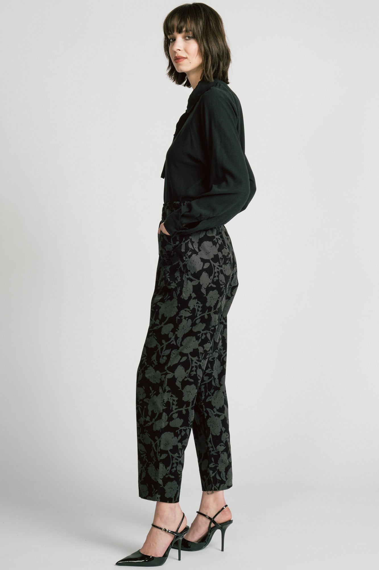 Side view of a woman wearing the Orion Pants by Allison Wonderland, relaxed fitting pants with an elastic waist and pockets in a dark Floral print on fine wale corduroy. She is wearing them with a black blouse and standing in front of a white background. 