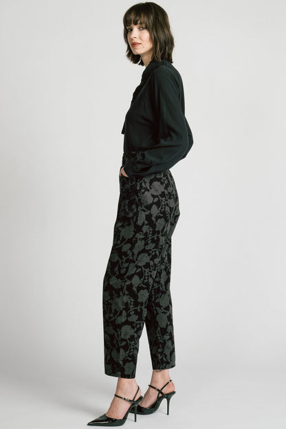 Side view of a woman wearing the Orion Pants by Allison Wonderland, relaxed fitting pants with an elastic waist and pockets in a dark Floral print on fine wale corduroy. She is wearing them with a black blouse and standing in front of a white background. 