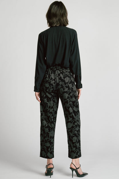 Back view of a woman wearing the Orion Pants by Allison Wonderland, relaxed fitting pants with an elastic waist and pockets in a dark Floral print on fine wale corduroy. She is wearing them with a black blouse and standing in front of a white background. 