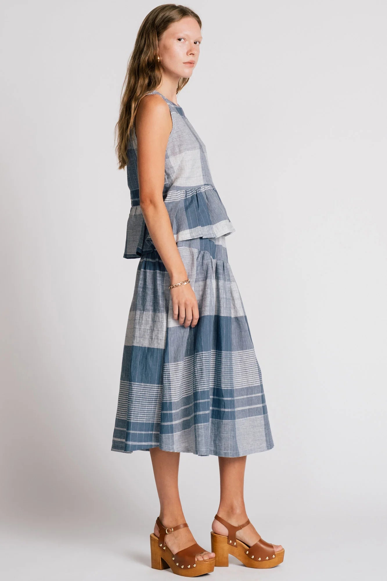 Side view of a woman wearing the Osprey Blouse by Allison Wonderland in Blue Plaid, a sleeveless top with a peplum both front and back and a cross-over back. She is wearing it with the matching Pelican Skirt and standing in front of a plain background. 

