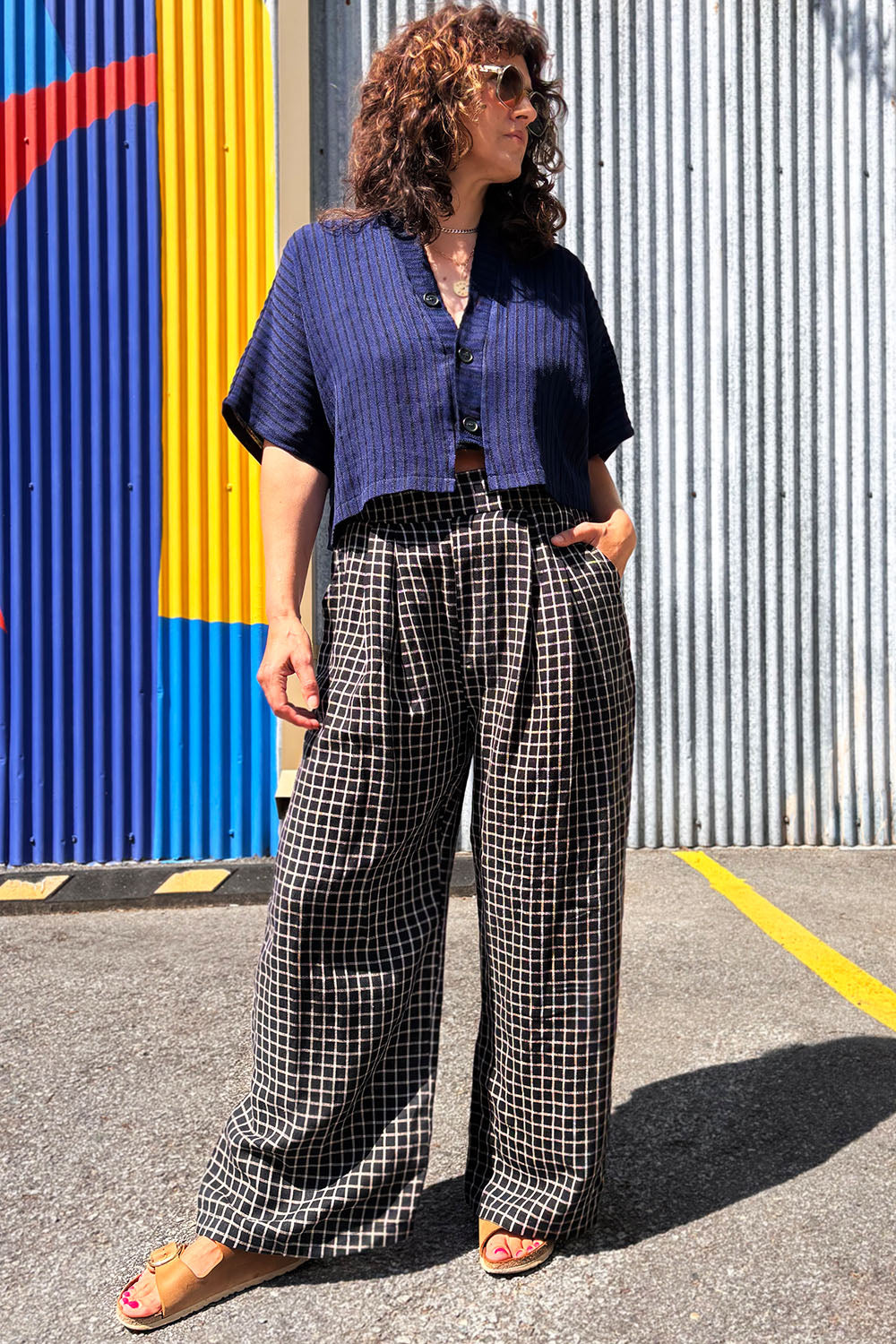 Woman wearing the Pip Jacket by Allison Wonderland in Navy/Black Stripe, with checkered pants standing against a colourful wall.