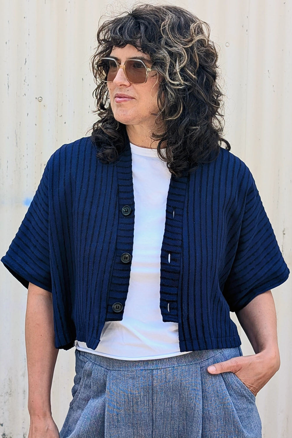 Woman wearing the Pip Jacket by Allison Wonderland in Navy/Black Stripe, with blue pants standing against a colourful wall.