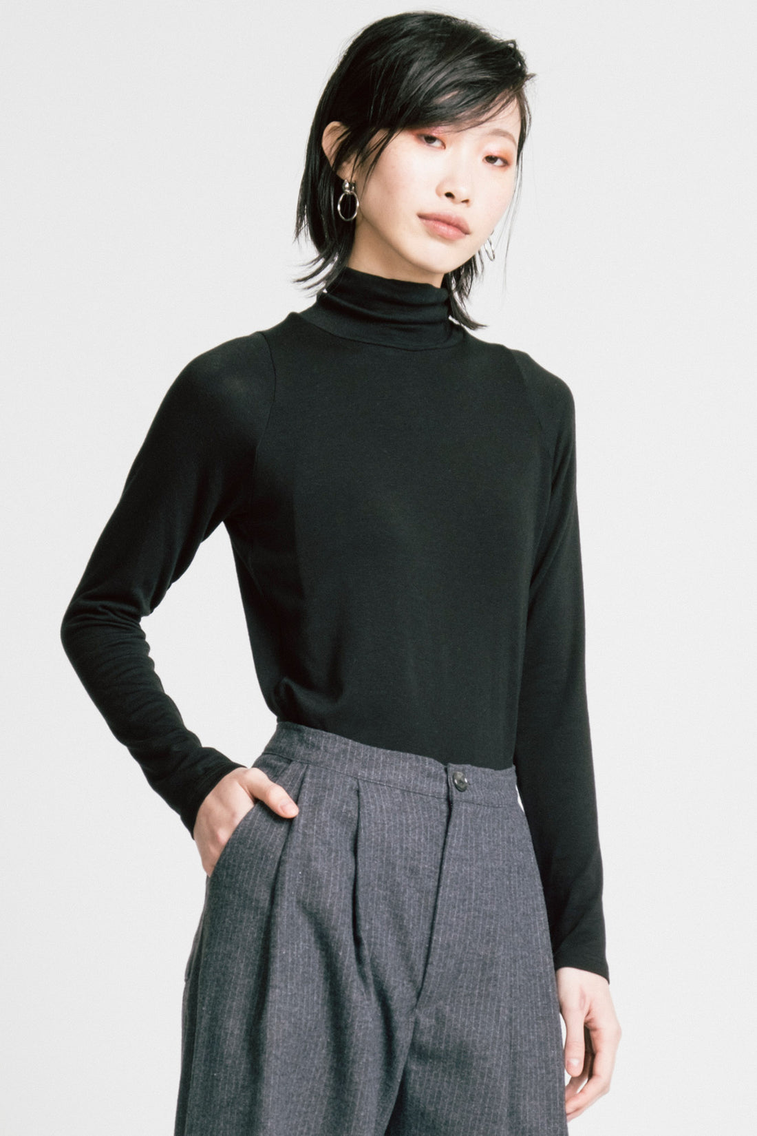 A woman wearing the Aches Turtleneck by Allison Wonderland in Black, a slim fitting classic turtleneck. She is wearing it with the  Memory Pants in Denim Stripe and standing in front of a white background. 
